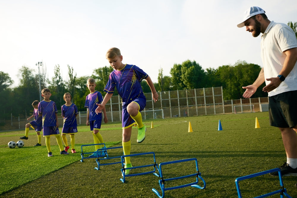 Young boys in purple and yellow soccer uniforms practice agility with blue hurdles on a grassy field. A coach in a white cap encourages them.