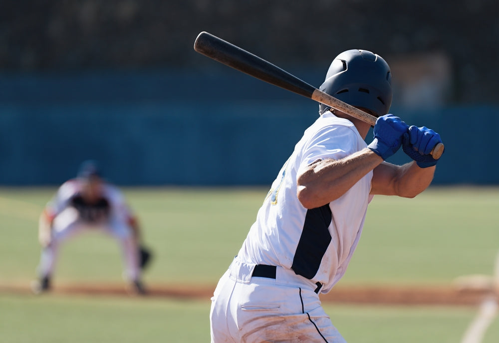 Baseball player in white uniform and helmet poised to bat, with a focused stance. Pitcher in blurred background, highlighting a tense game moment.